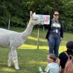 A librarian reading a book in an field while a family and an alpaca listen.