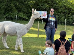 A librarian reading a book in an field while a family and an alpaca listen.